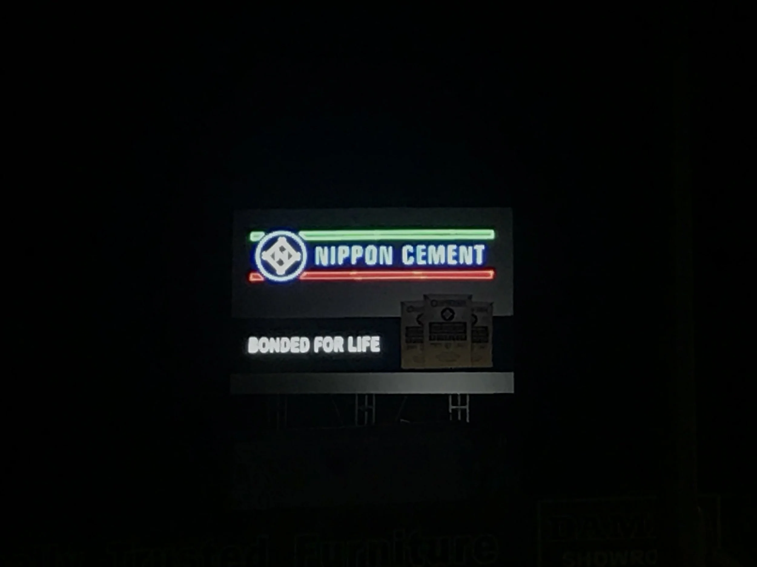 A neon sign for Nippon Cement with a logo, the words 'Nippon Cement,' and the phrase 'Dedicated for Life' below it, illuminated against a dark background.