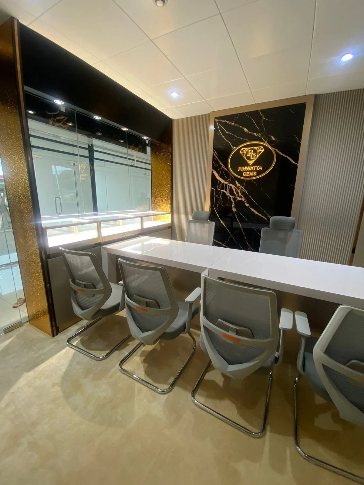 A modern office conference room with a white table, six gray office chairs, and a black marble wall featuring a gold logo that reads 'PINAWATTA GEMS' with a diamond icon.