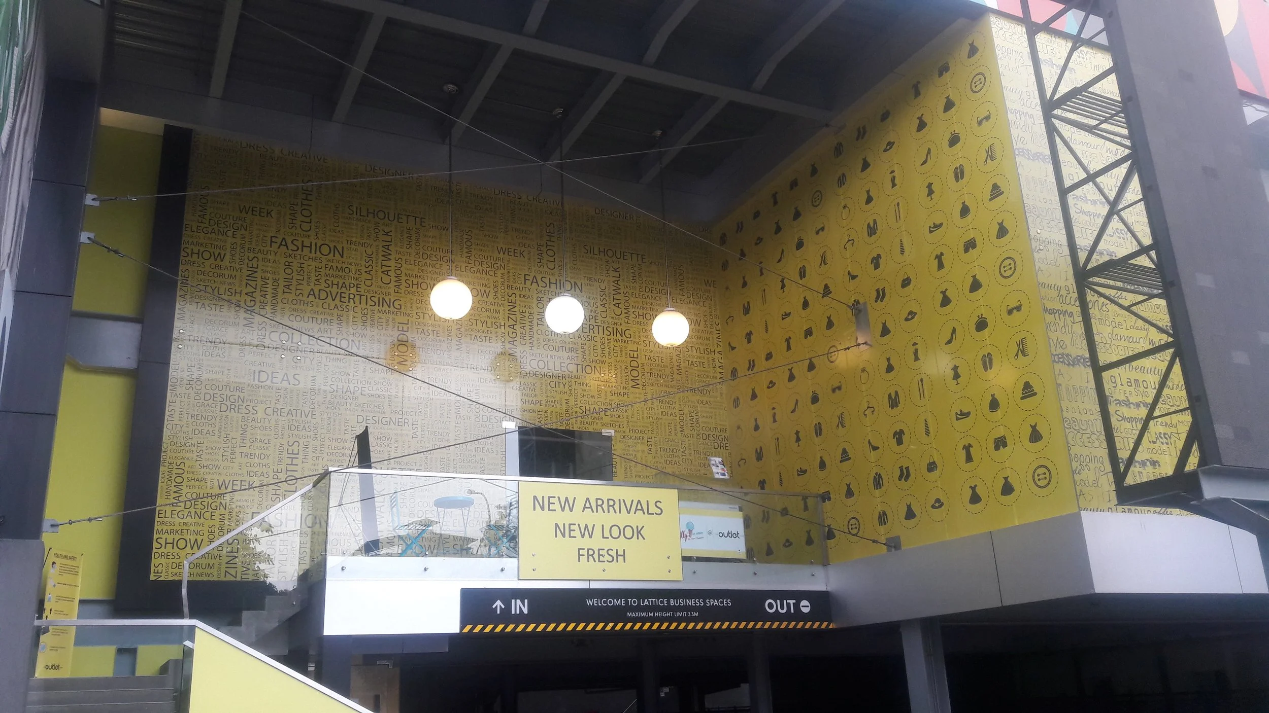 Interior of a shopping mall or retail space featuring bold yellow wall with icons and words related to fashion and shopping. A sign indicates new arrivals and a welcome message to Lattice Business Spaces, with entry and exit directions.