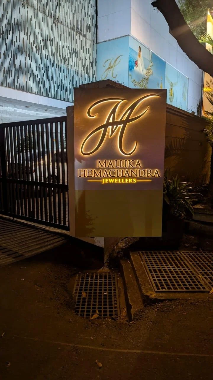 A sign for Mallika Hemachandra Jewellers illuminated at night, with a building and plants in the background.