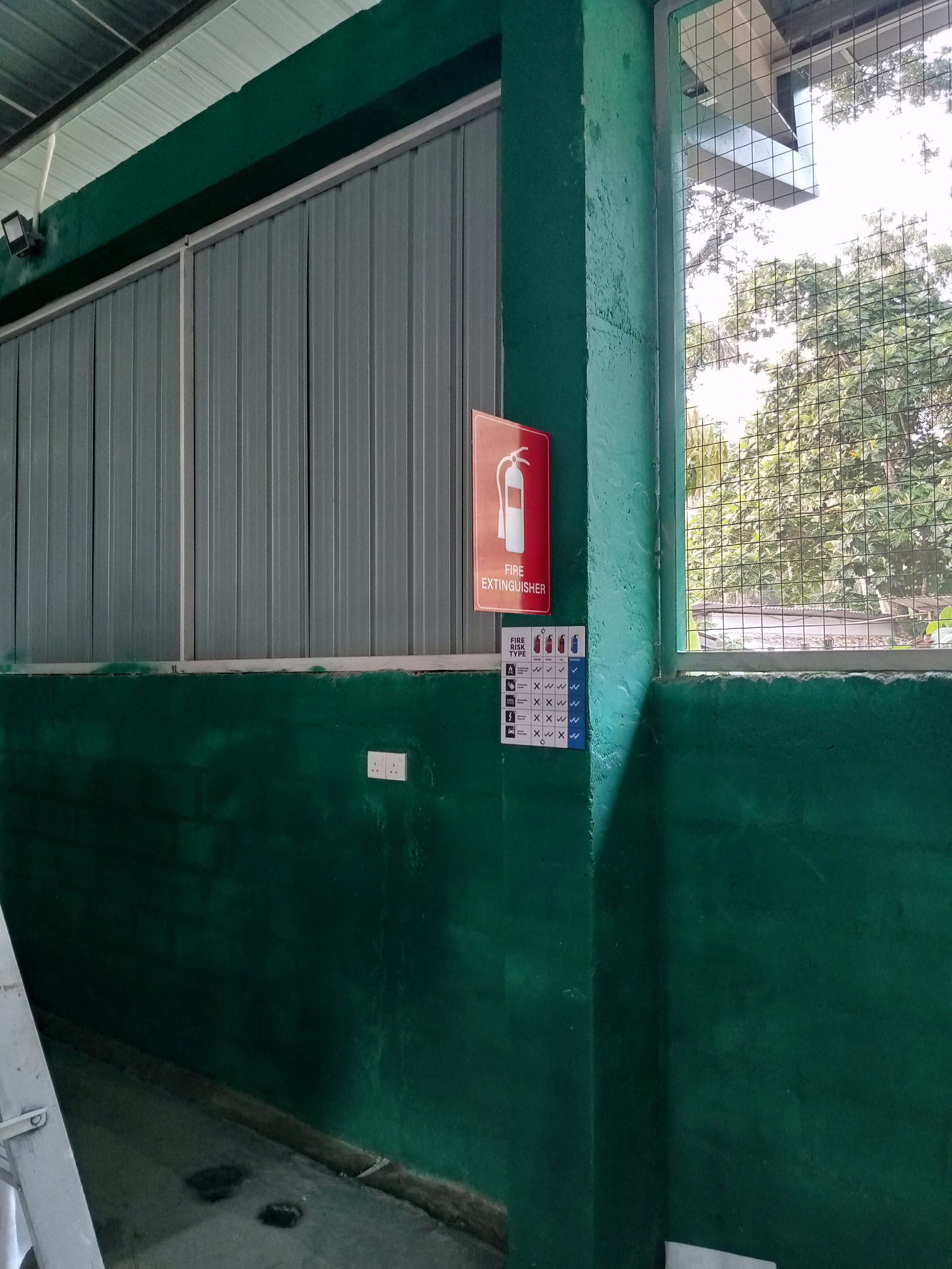 Indoor green-painted wall with a fire extinguisher sign and a wire mesh window showing trees outside.