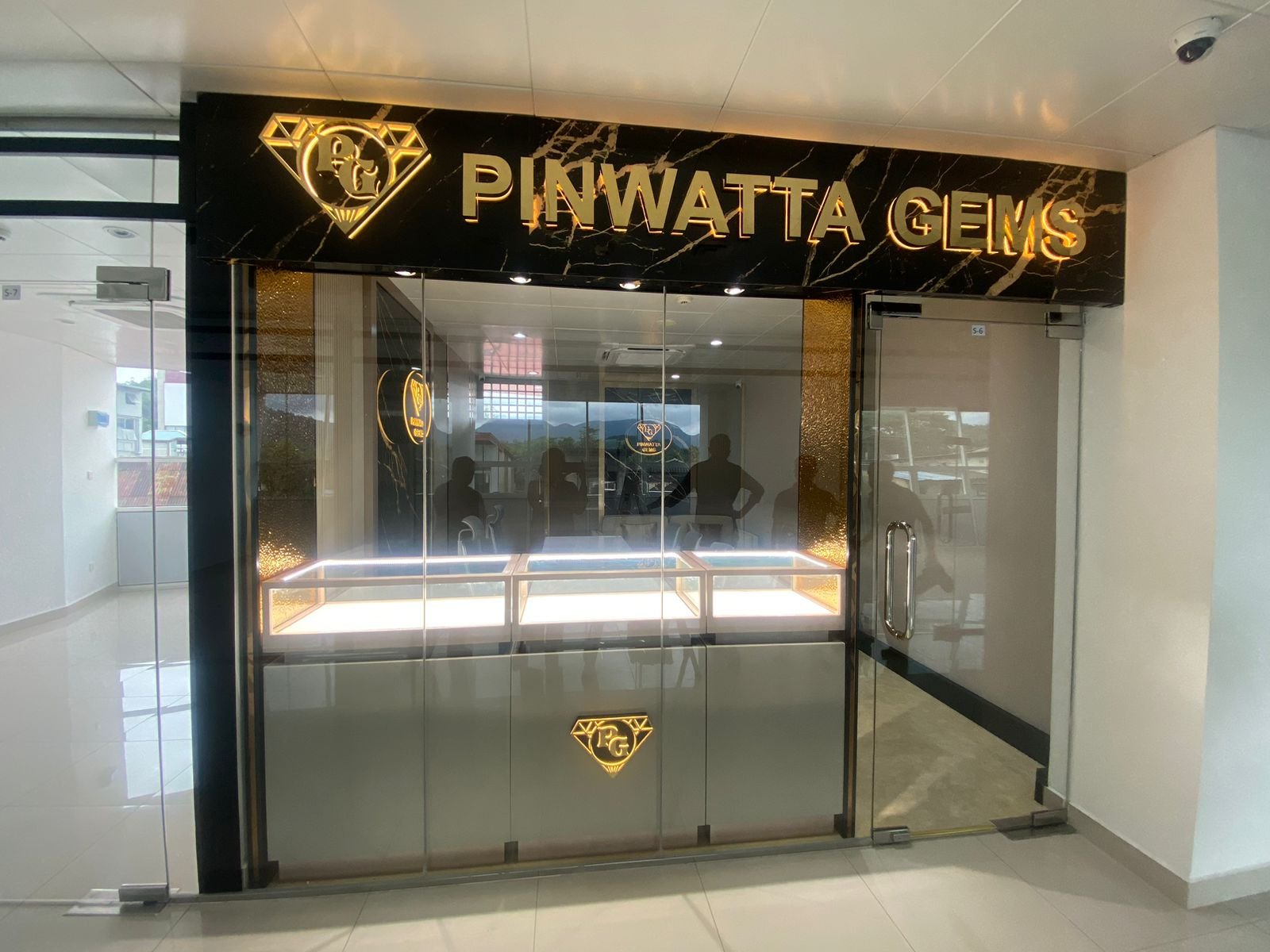  storefront with black marble signage featuring a diamond-shaped logo and the text 'PINWATTA GEMS' in gold illuminated letters, glass doors with reflections of people inside, and jewelry display cases.