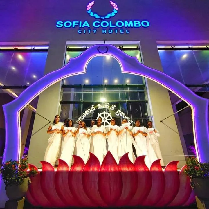 Seven women dressed in white sarees standing on a stage in front of the Sofia Colombo City Hotel, with a purple-lit decorative arch and a lotus-shaped base, at night.