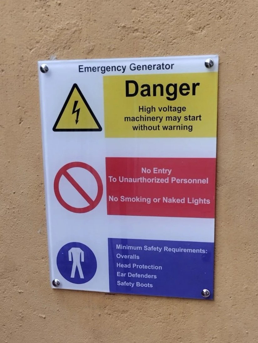 Warning sign for an emergency generator with instructions: danger high voltage machinery may start without warning, no entry to unauthorized personnel, no smoking or naked lights, and safety requirements like overalls, head protection, ear defenders, and safety boots.