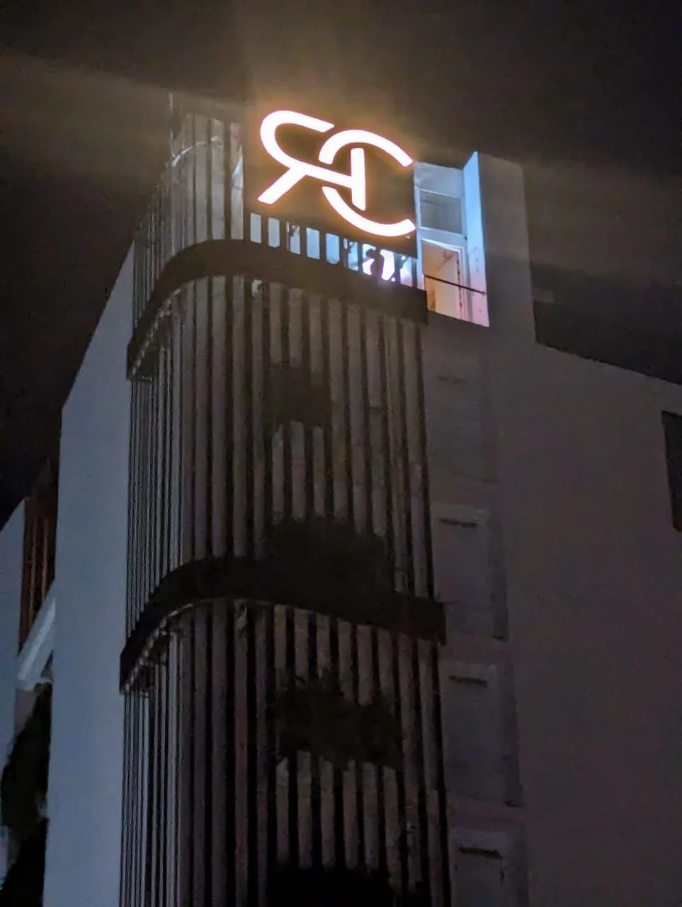 Nighttime view of a building with a illuminated sign displaying initials or a logo on top, and a spiral staircase on the exterior.