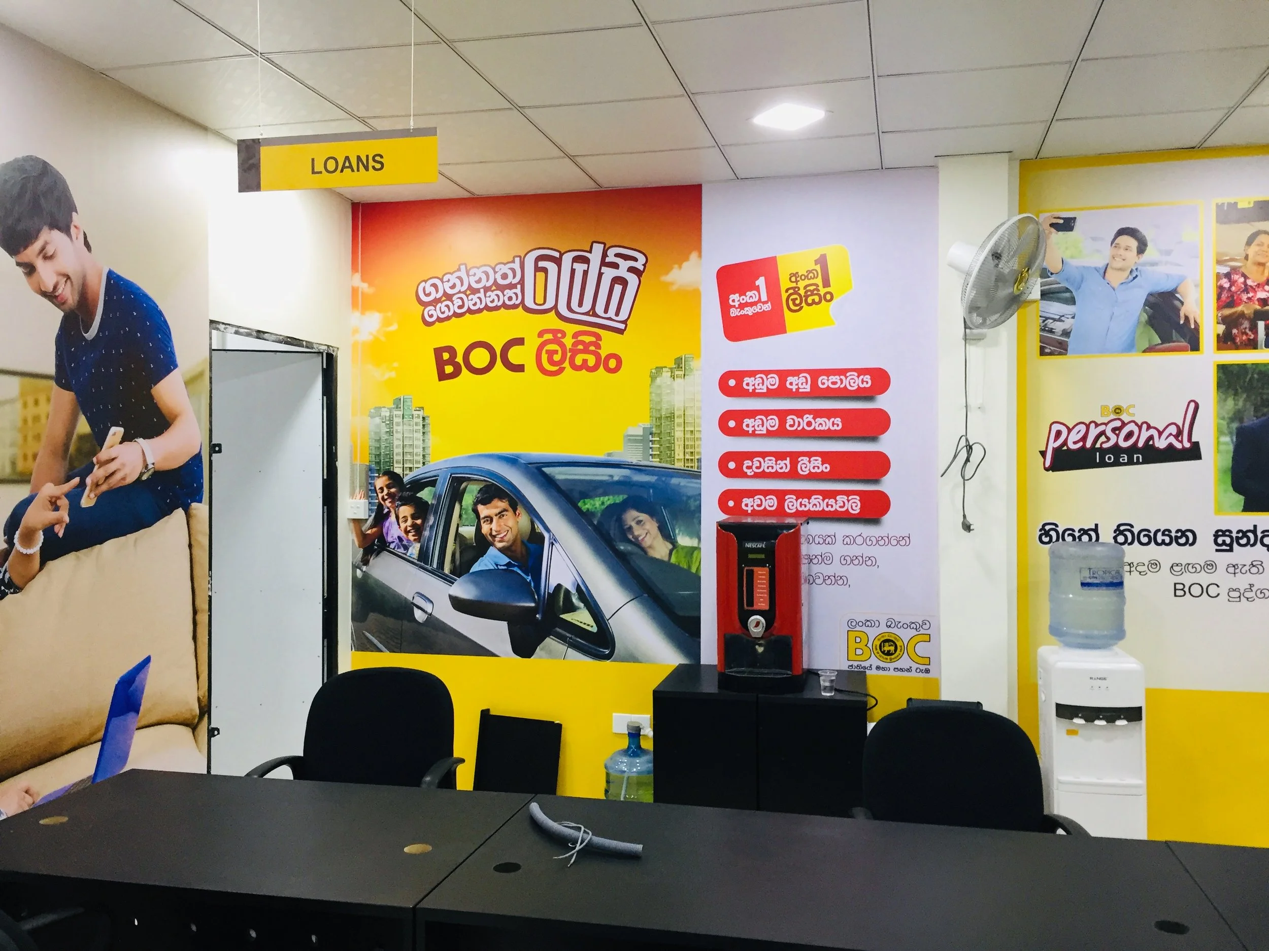 Wall advertisements showing people smiling and taking selfies, with a yellow sign labeled 'LOANS' hanging from the ceiling in a brightly lit room.