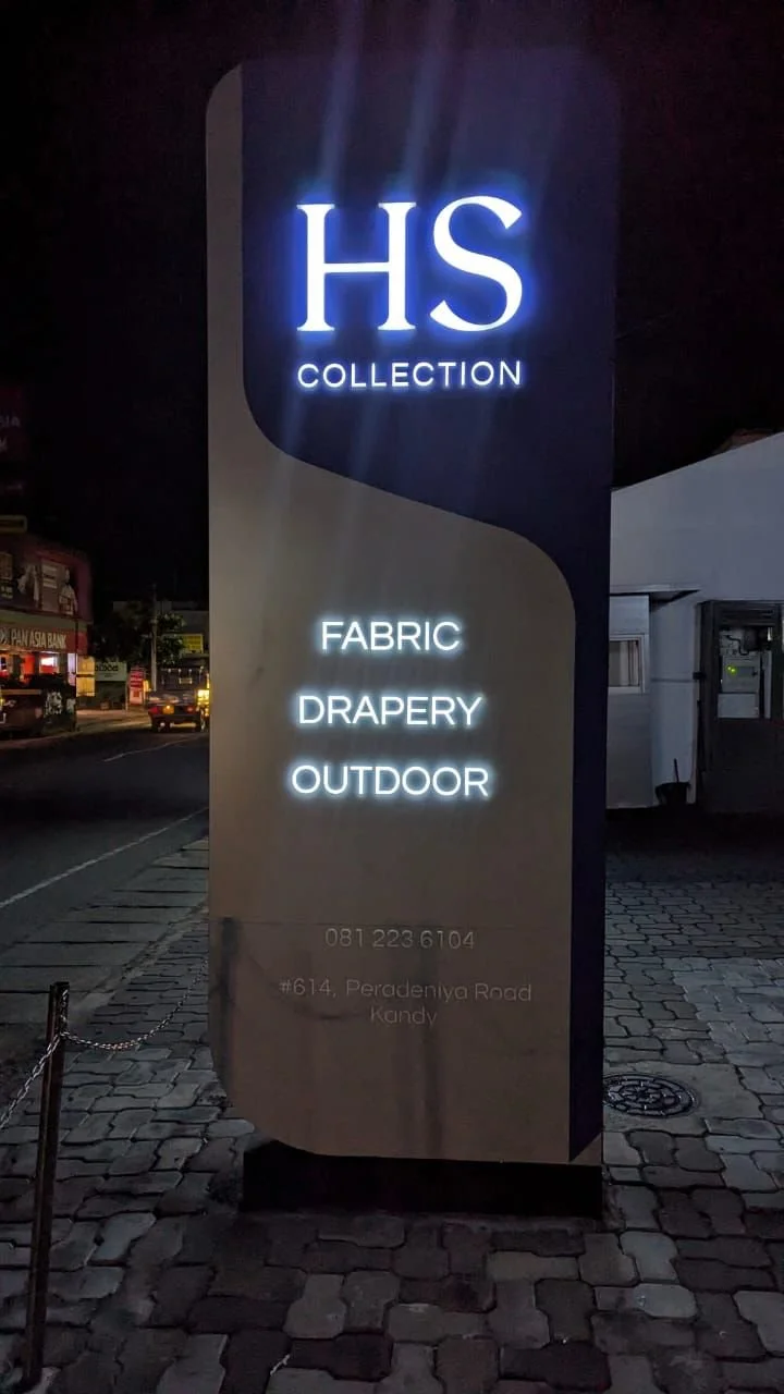 Illuminated sign for HS Collection featuring fabric, drapery, and outdoor products, with contact number 081 223 6104 and address #614, Peradeniya Road, Kandy, at night.