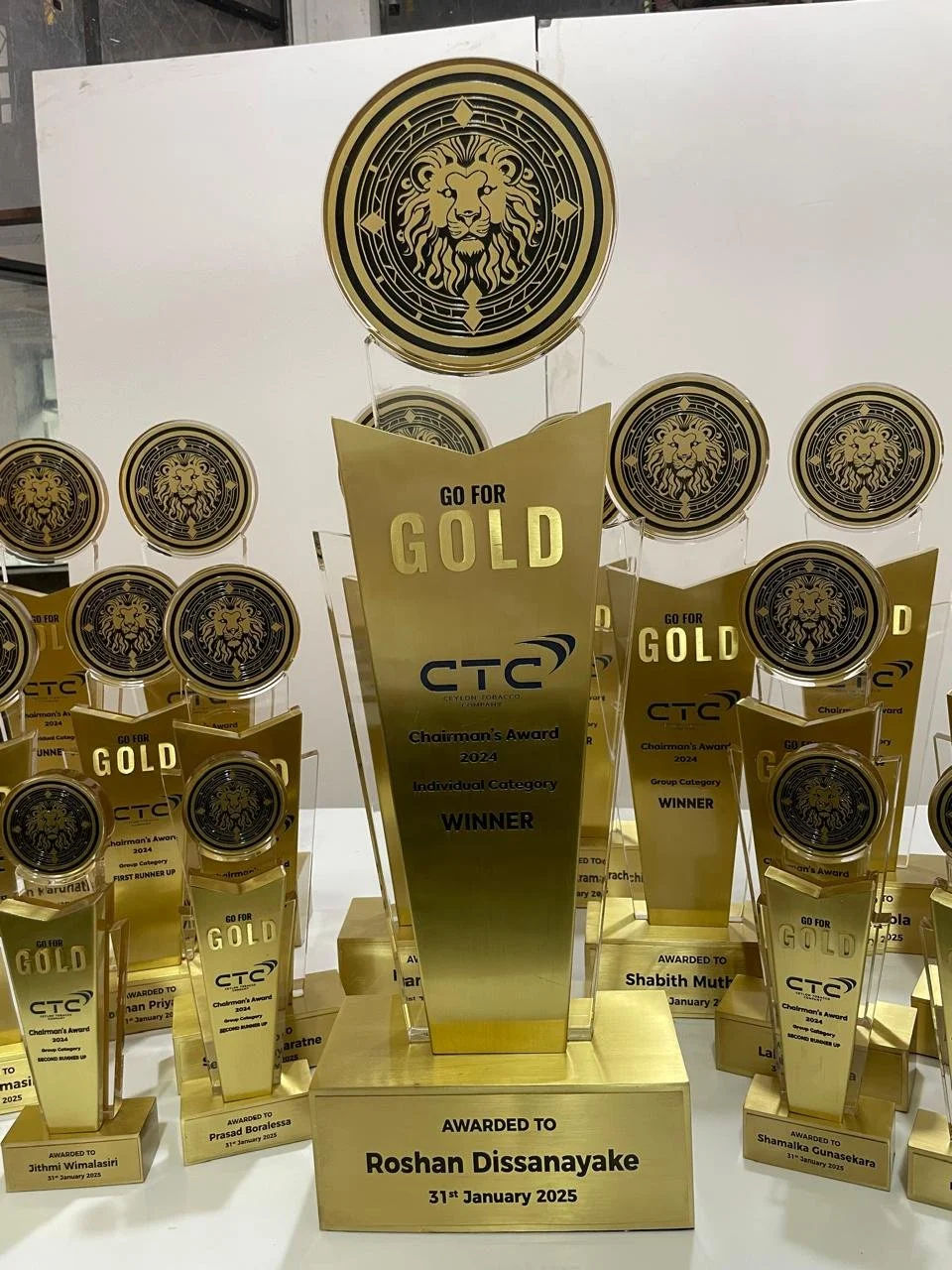 Collection of gold-colored award trophies with a lion logo, awarded to Roshan Dissanayake on January 31, 2025, for excellence in the CTI Chairman's Award 2024, with similar trophies and a lion emblem in the background.