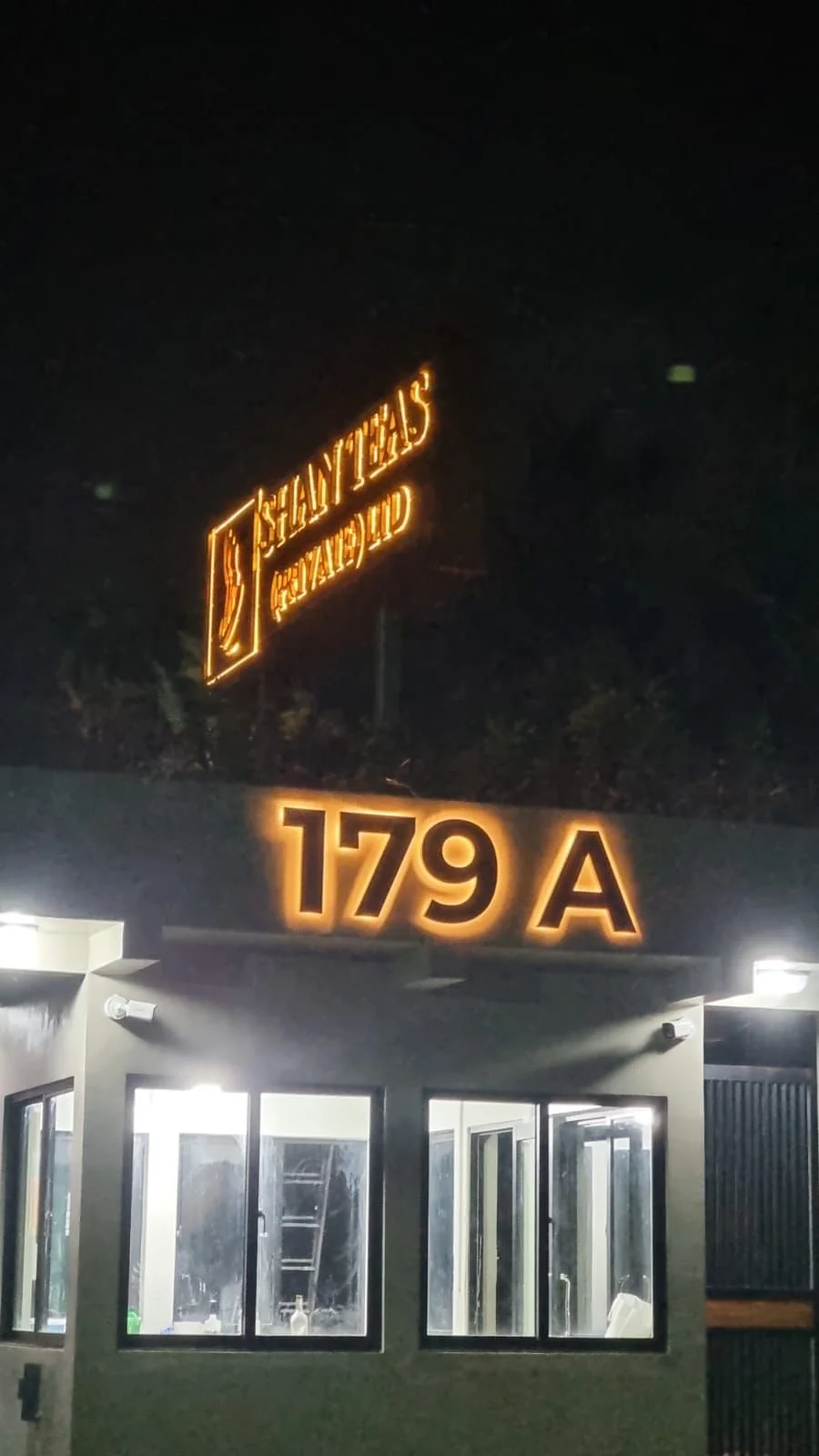 Night view of a building entrance with illuminated signs reading '179 A' and 'Shay Tesh Original,' with large windows showing the inside of the building.