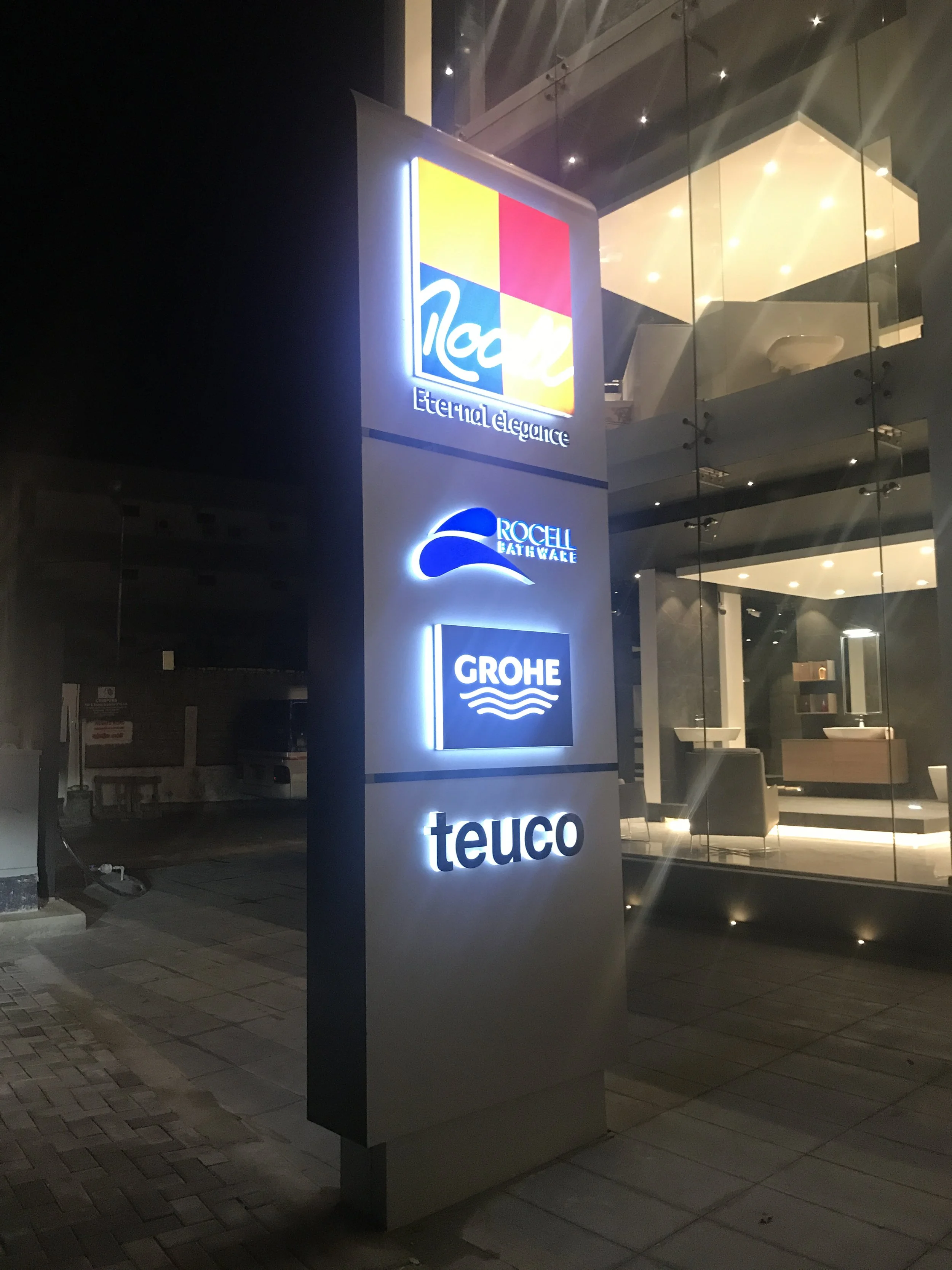 A tall illuminated sign with logos of Nola, Eterna elegance, Rocell Bathware, Grohe, and Teuco outside a modern building at night.