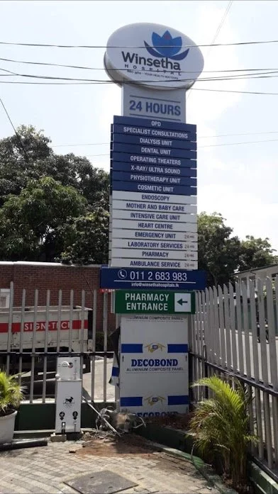 Signboard for Winsetha Hospital featuring services like dialysis, dental, physiotherapy, endoscopy, pharmacy, ambulance, and more, with a pharmacy entrance arrow and contact number.