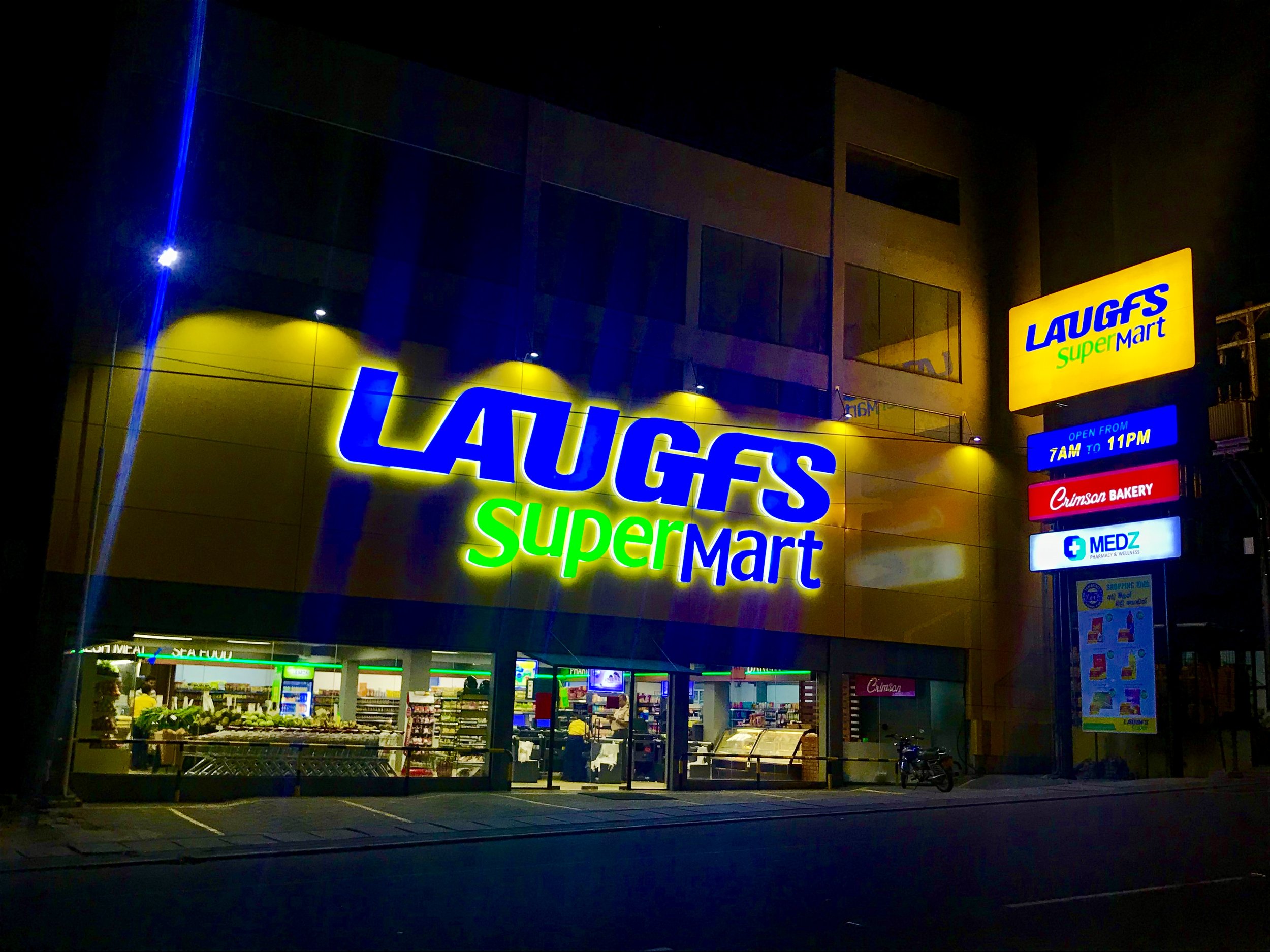 Night view of a Laugs Supermarket with illuminated signs, including purple, yellow, green, and red, and a visible storefront with groceries and shoppers inside.