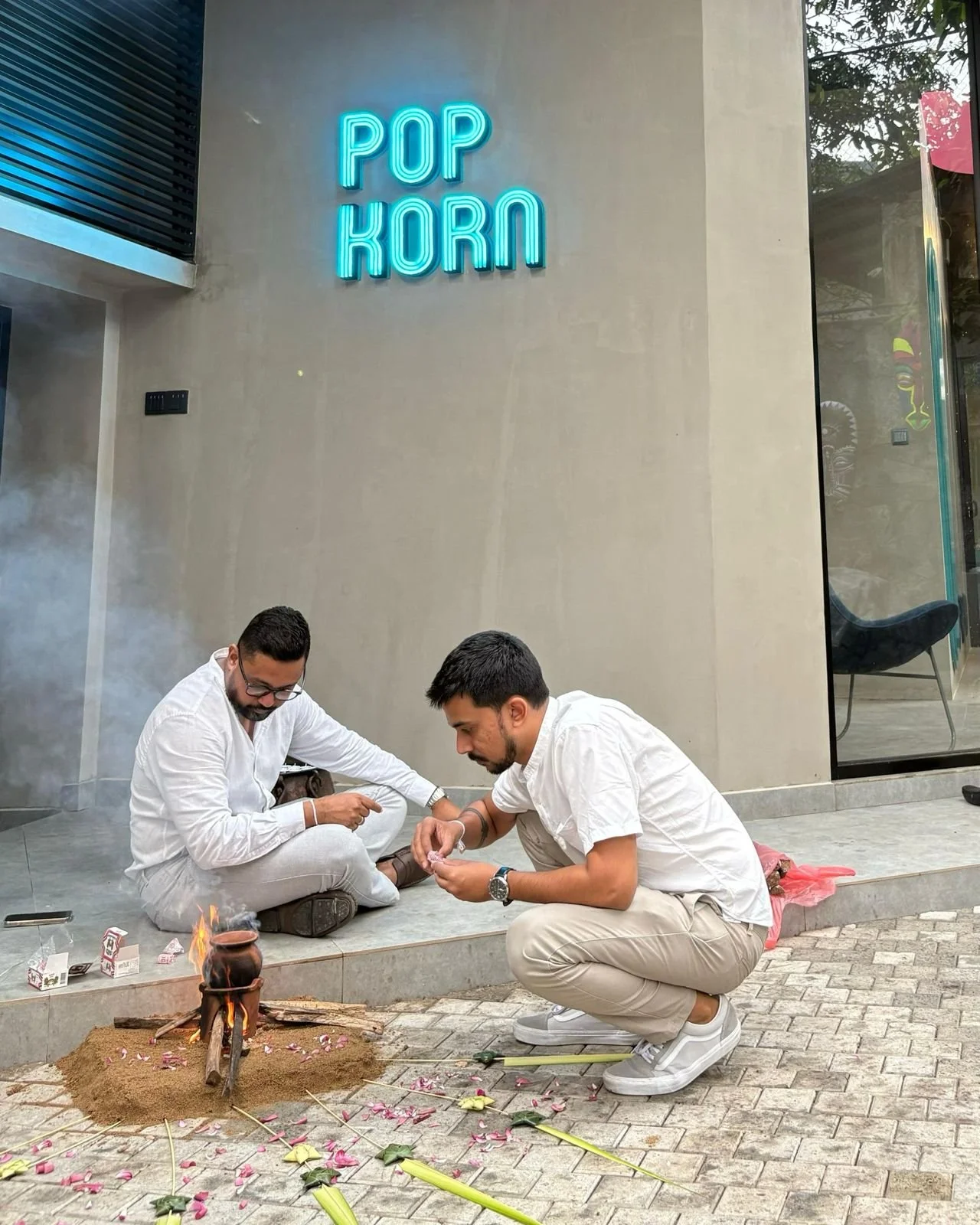 Two men sitting on the sidewalk in front of a building with a neon sign reading 'POP KORN'. They are preparing to perform a traditional fire ritual with small clay pots, flowers, and small fires on the ground. The men are wearing white shirts and lig