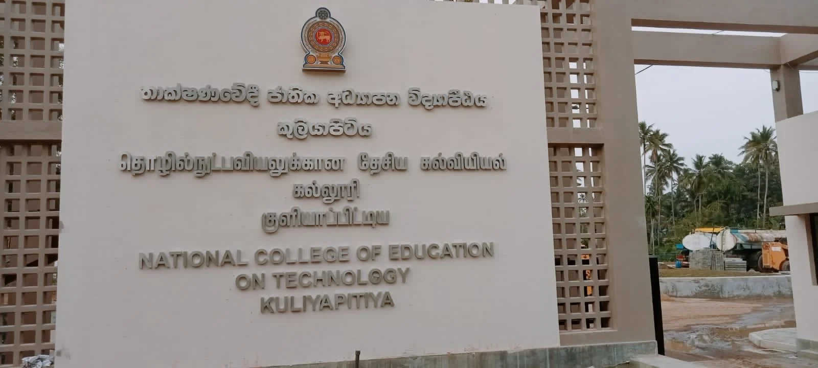 Entrance sign for the National College of Education on Technology in Kuliyapitiya, Sri Lanka, with coconut trees and a construction vehicle in the background.
