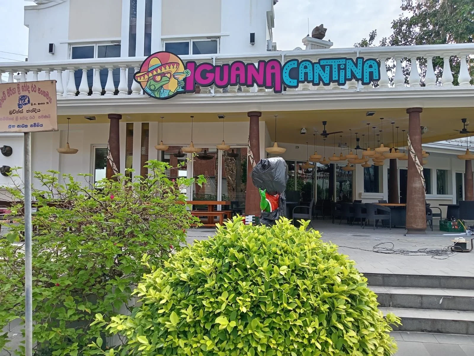 Exterior of a building with a sign that reads 'Guanacatina' with a cartoon frog wearing sunglasses and a sombrero, holding a cocktail. There are green bushes in the foreground and some setup equipment in front of the building.