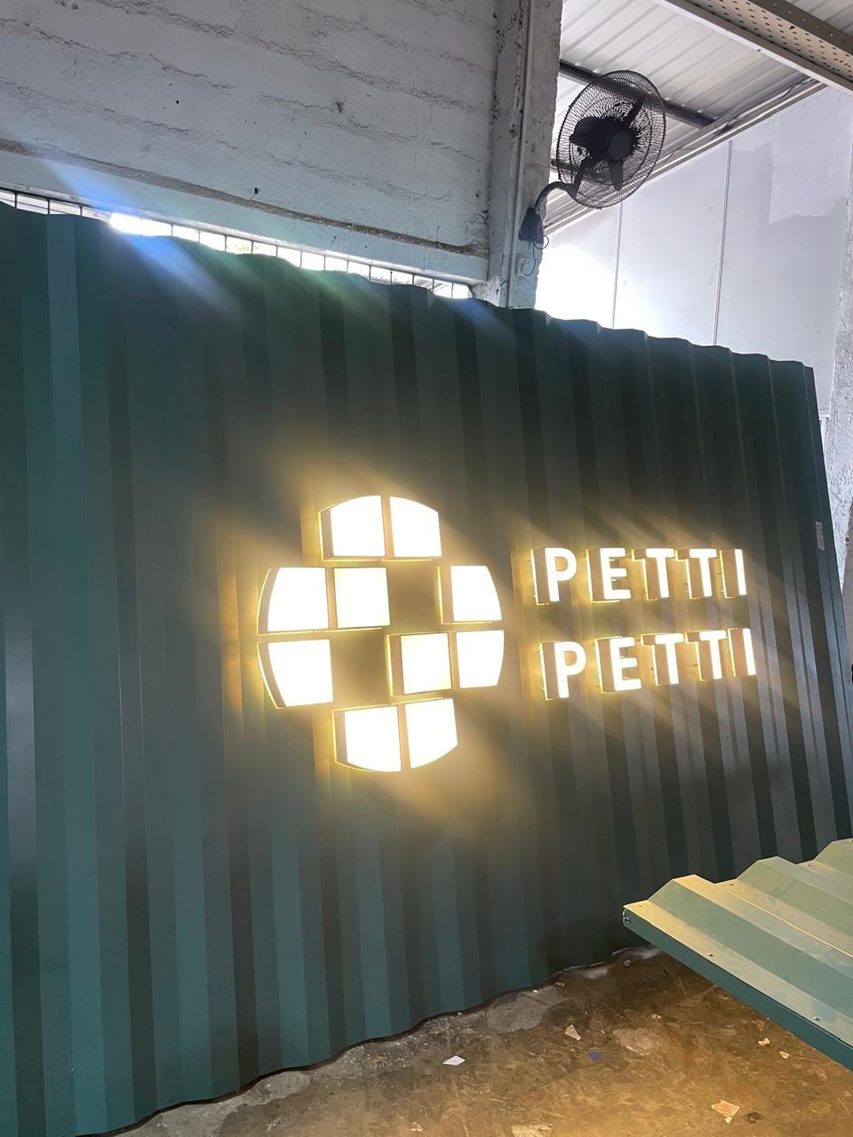 A illuminated sign displaying the text "PETTI PETTI" next to a cross logo, mounted on a dark green corrugated metal wall, with a small ceiling fan and a concrete ceiling visible in the background.