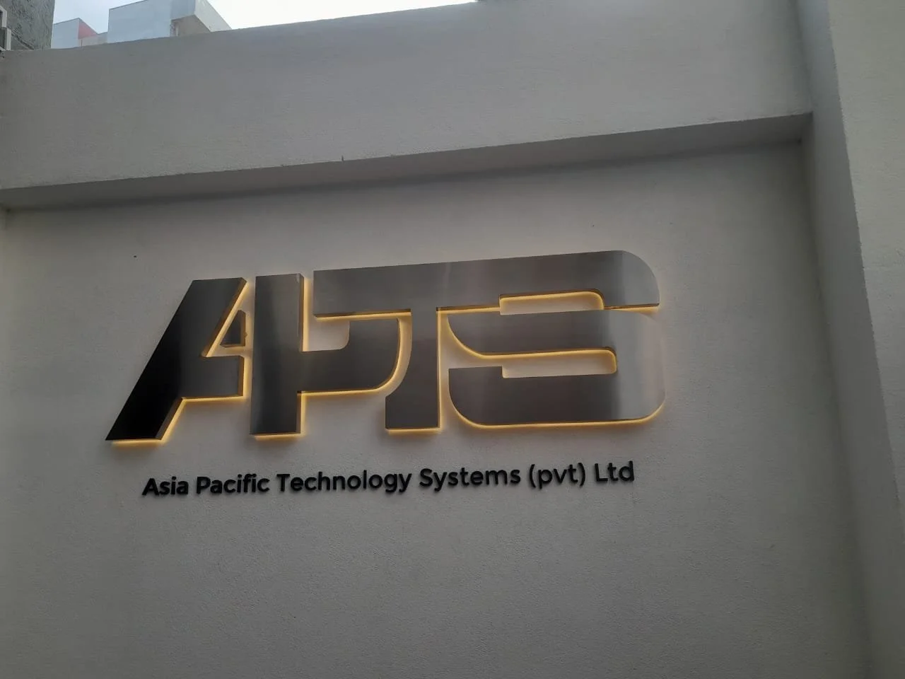Signboard displaying the logo and name of Asia Pacific Technology Systems (pvt) Ltd, with illuminated letters on a white wall.