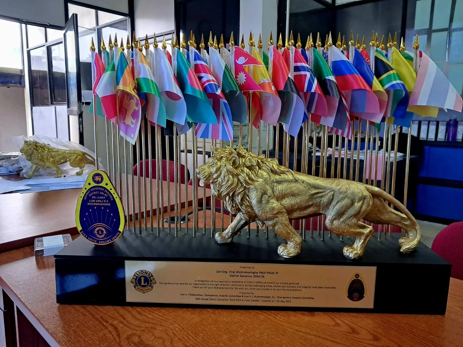 A decorative display featuring a group of small flags from various countries, a golden lion figurine, and a Lions Club emblem. The display is set on a table in an indoor setting with chairs and windows in the background.