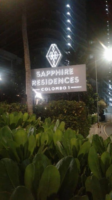 Nighttime view of a lit sign for Sapphire Residences Colombo, with plant bushes in the foreground and a tall building with lights behind.