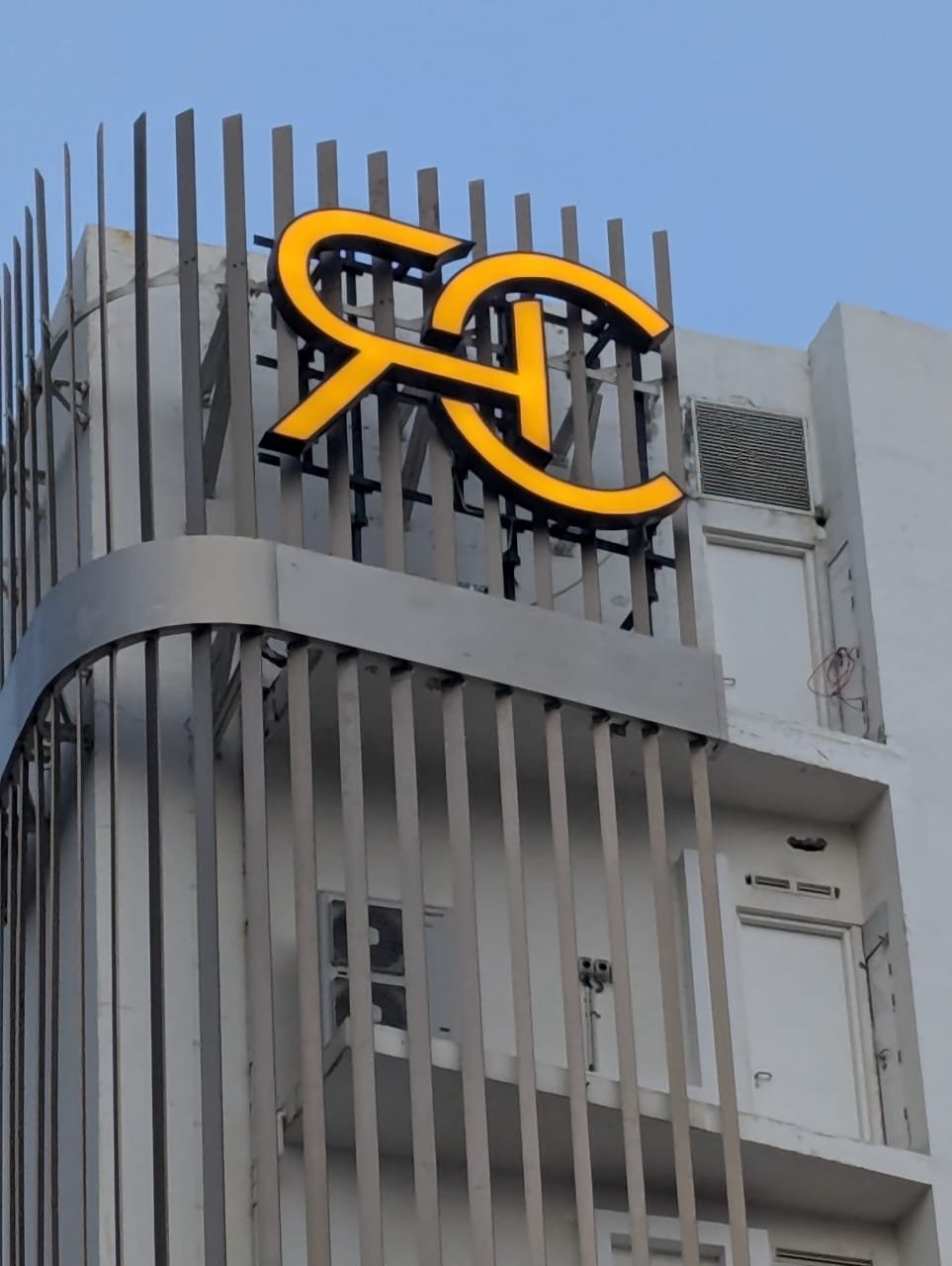 Hotel sign with illuminated logo of a yellow letter 'R' with a black outline, mounted on a metal framework on a white building facade with vertical metal bars.