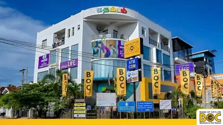 A white multi-story commercial building with colorful signage, including BOC Bank signs, trees, and a blue sky.