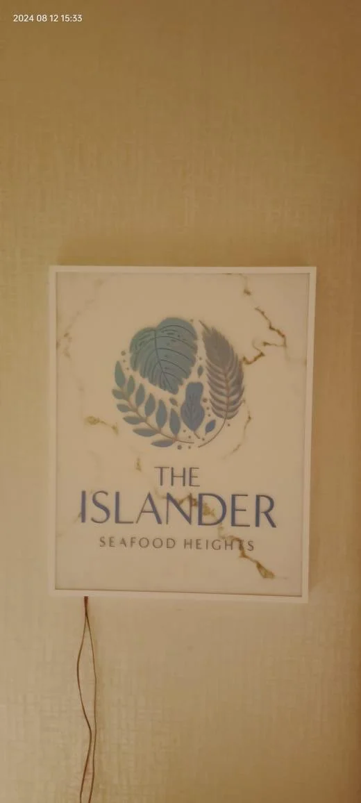 A sign with the text "The Islander Seafood Heights" featuring a logo with ocean leaves, mounted on a beige wall with visible cracks and a loose wire hanging below.