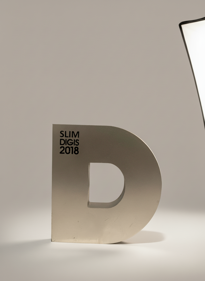 Silver letter D with black text 'SLIM DIGIS 2018' on a white surface, lit by a studio light in the upper right corner.