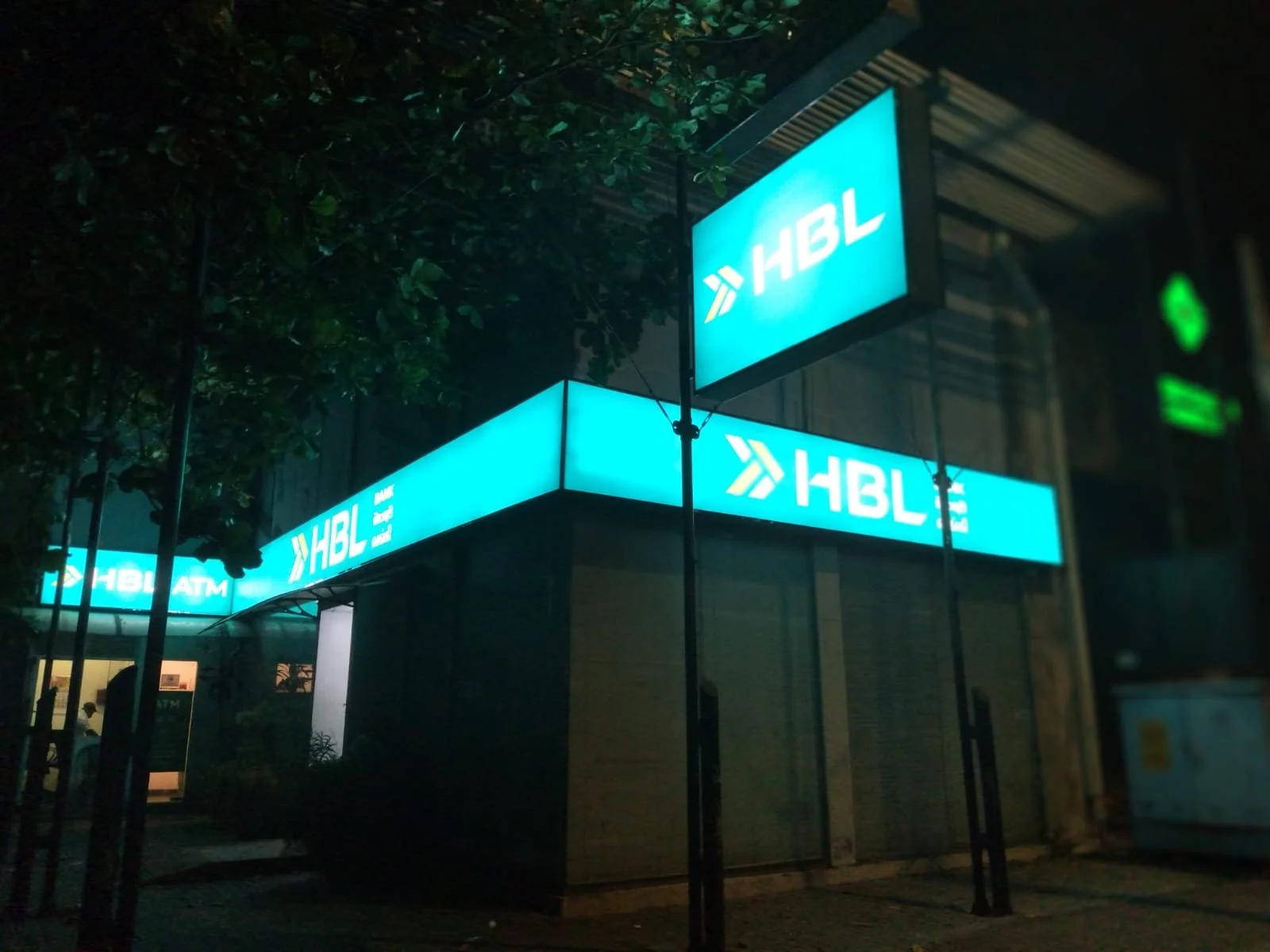 Nighttime scene of a building with illuminated blue HBL signages, including an ATM, partially obscured by tree branches.