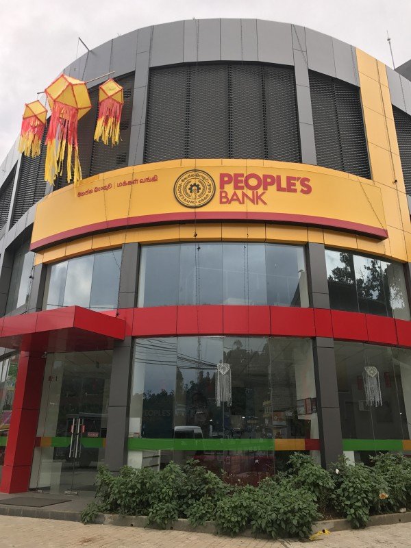 A modern building with a yellow sign reading "People's Bank" featuring the bank's logo and text in another language. The building has large glass windows and is decorated with colorful lanterns hanging outside.