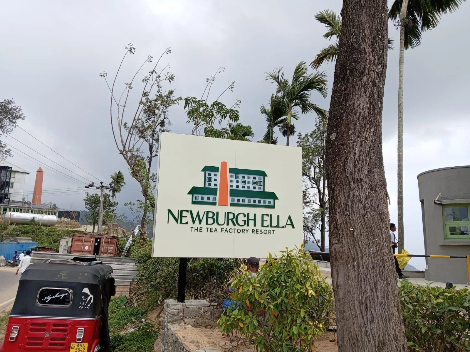 Sign for Newburgh Ella, The Tea Factory Resort, with trees and buildings in the background, and a three-wheeled auto rickshaw in the foreground.