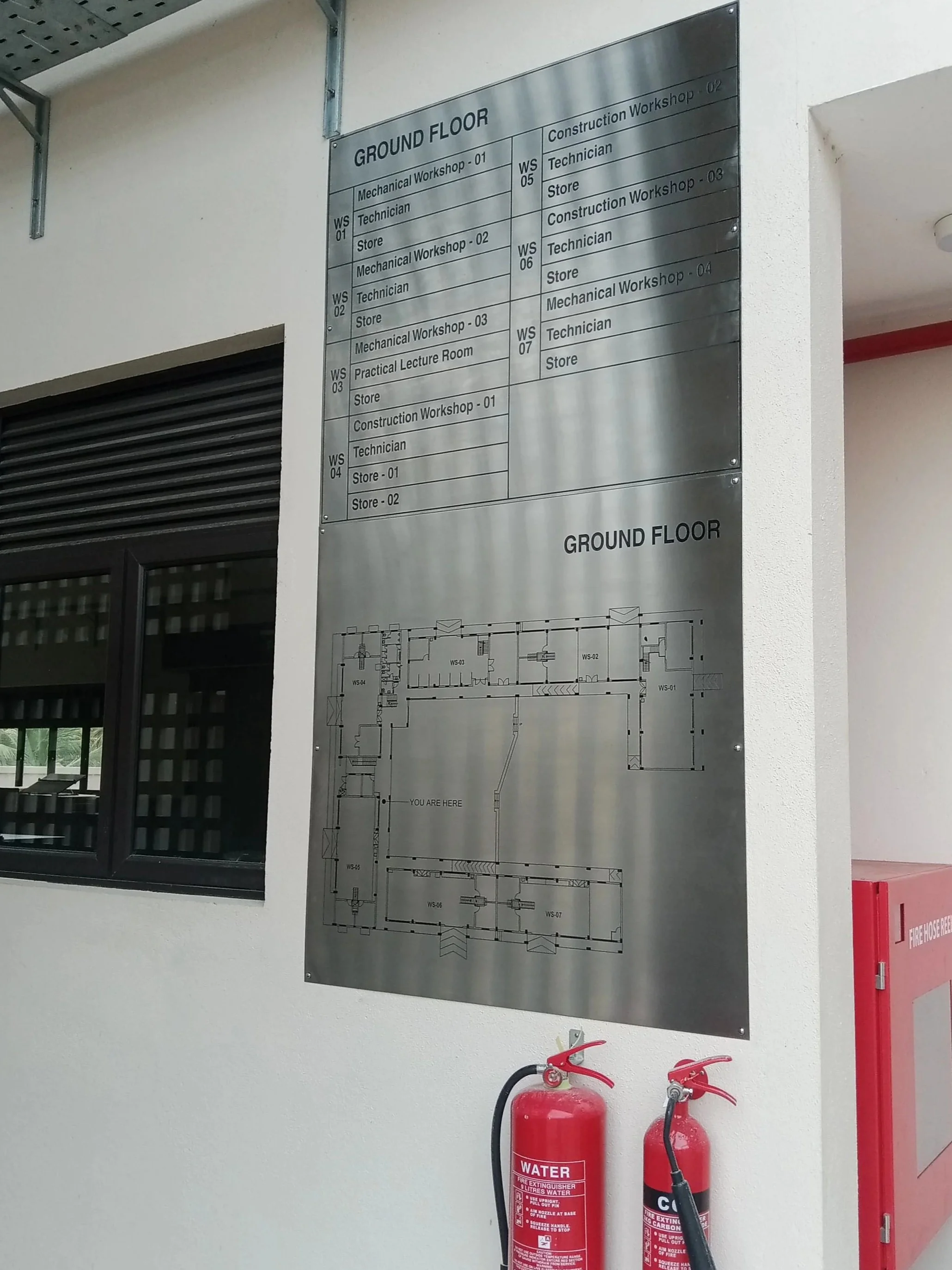 A large metallic sign charting the ground floor room layout and design, with a black-framed window to the left and two red fire extinguishers below the sign. The wall is white and there is a fire hose reel nearby.