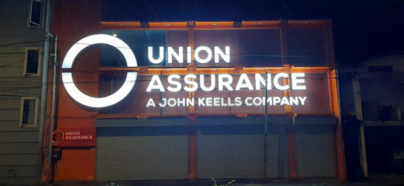 Nighttime exterior of a building with a large illuminated sign reading 'Union Assurance, A John Keells Company' and a logo featuring a circle with a gap.