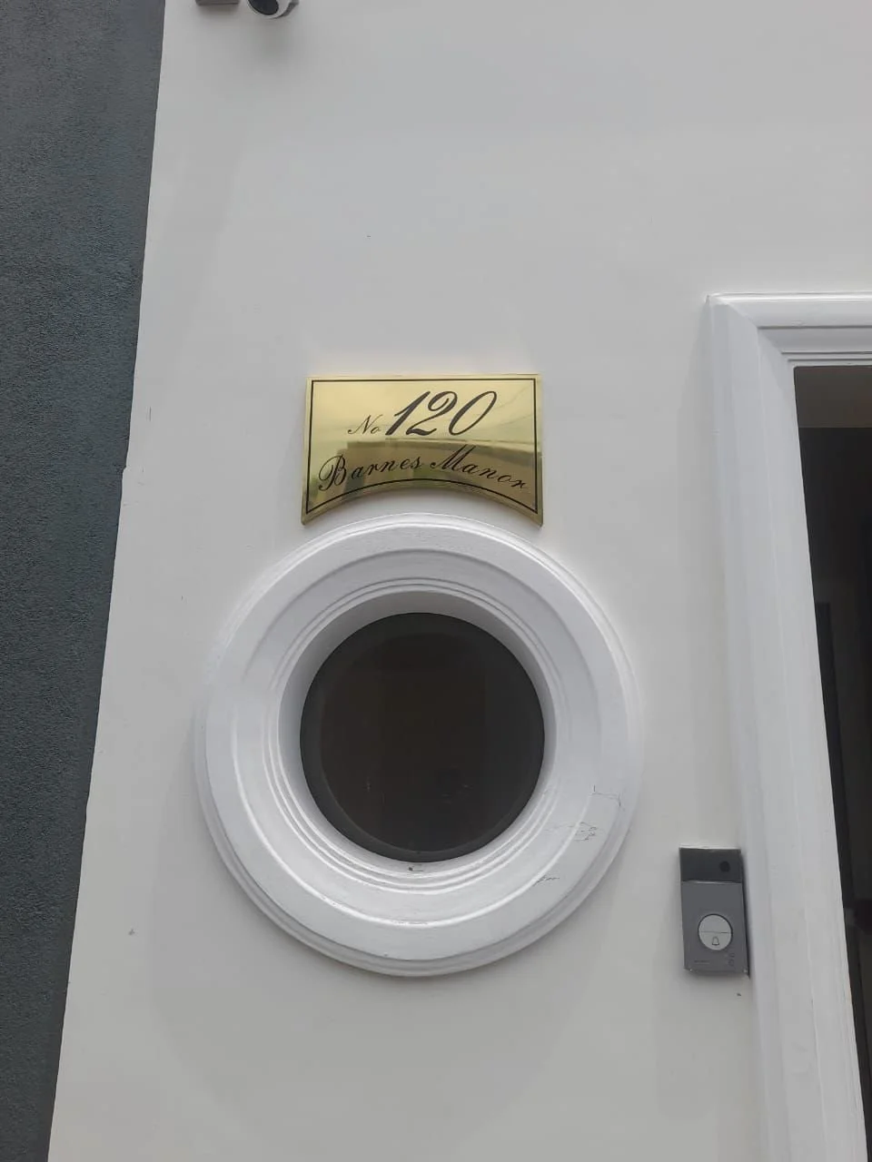 Gold house number plaque with black cursive text 'No 120 Barnes Manor' above a circular window with a white decorative frame on a white wall.