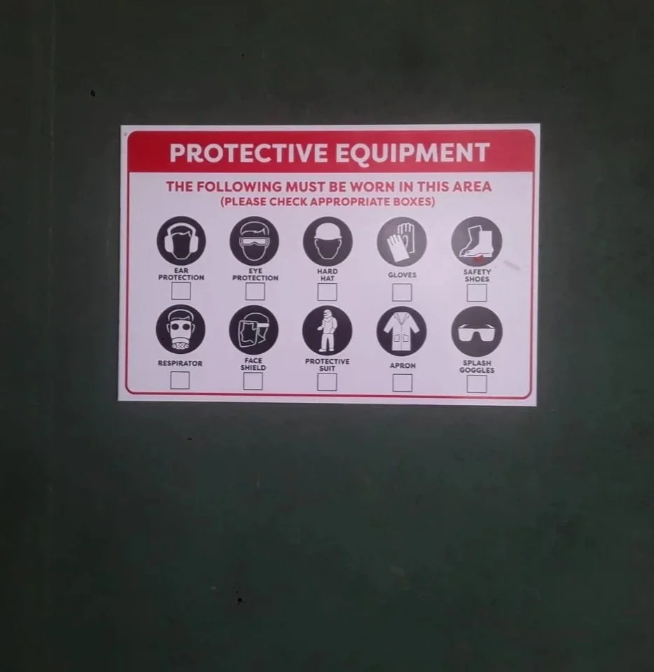 Sign listing required protective equipment including ear protection, eye protection, hard hat, gloves, safety shoes, respirator, face shield, protective suit, apron, and splash goggles.