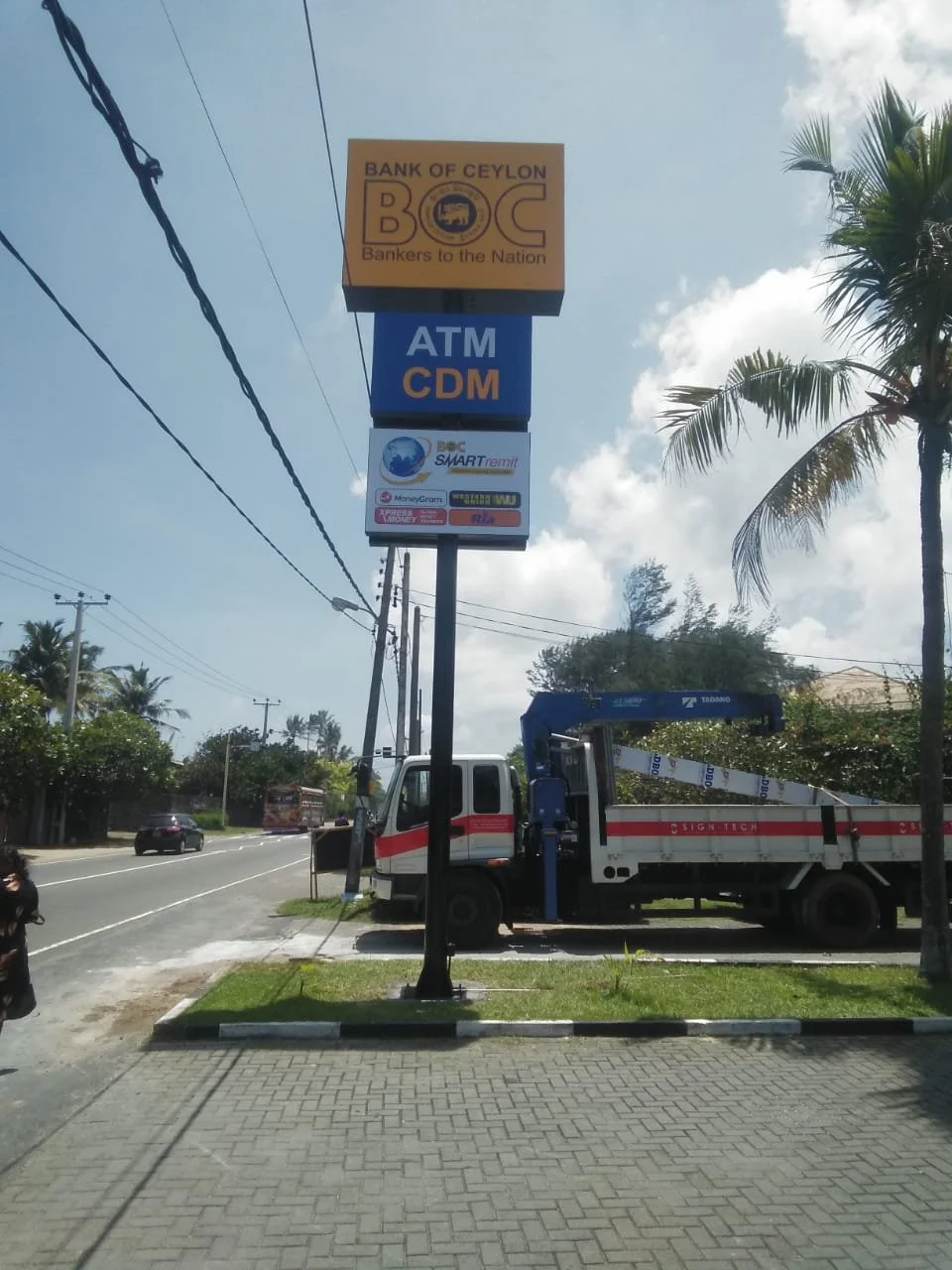 Signboard for Bank of Ceylon with advertisement for ATM CDM, and smaller logos for SMART Remit and other services, with a street scene including a truck and trees.