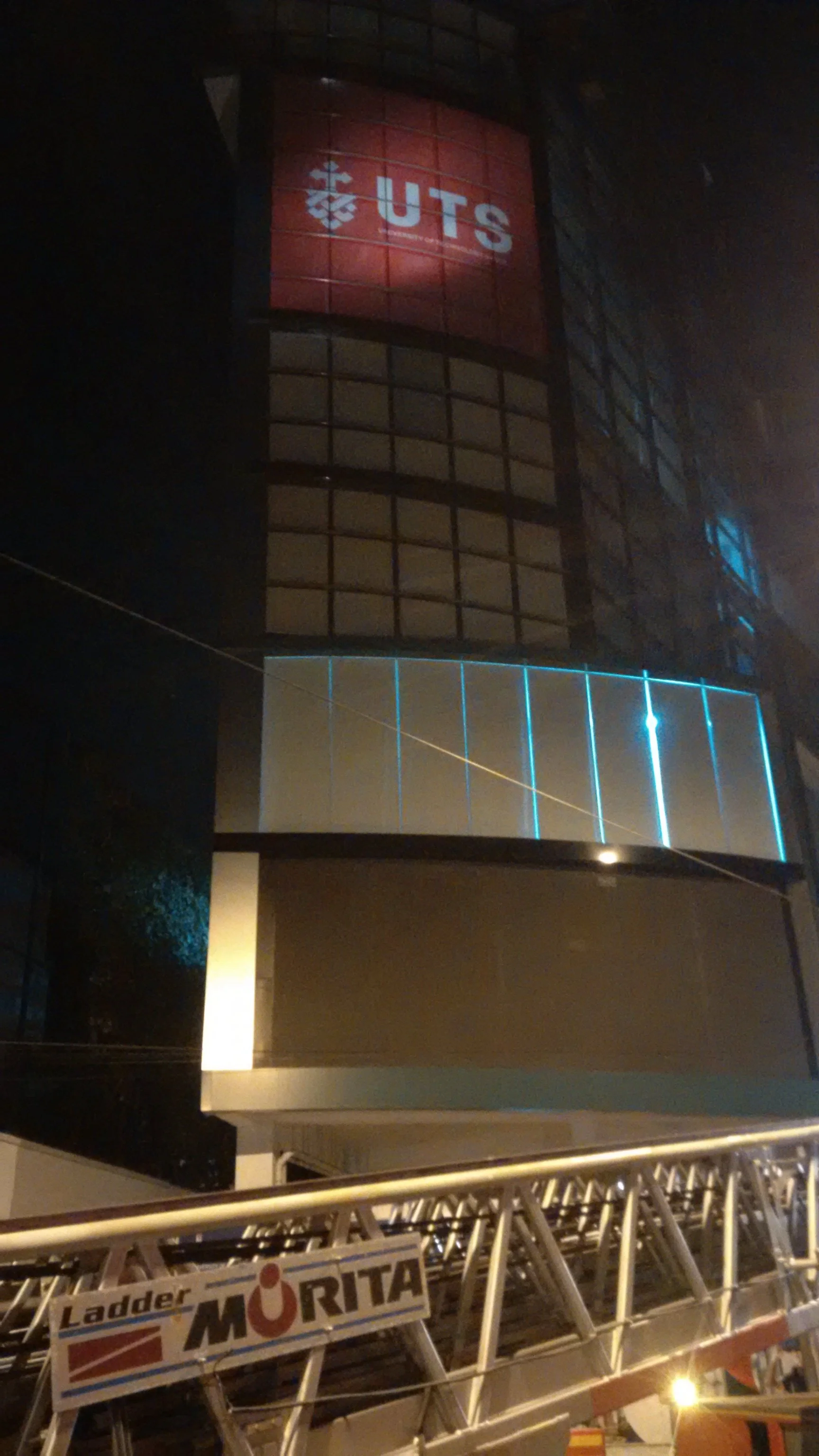 Nighttime image of a building with a red illuminated sign that reads 'UTS,' and a barricade with the word 'MORITA' at the bottom.