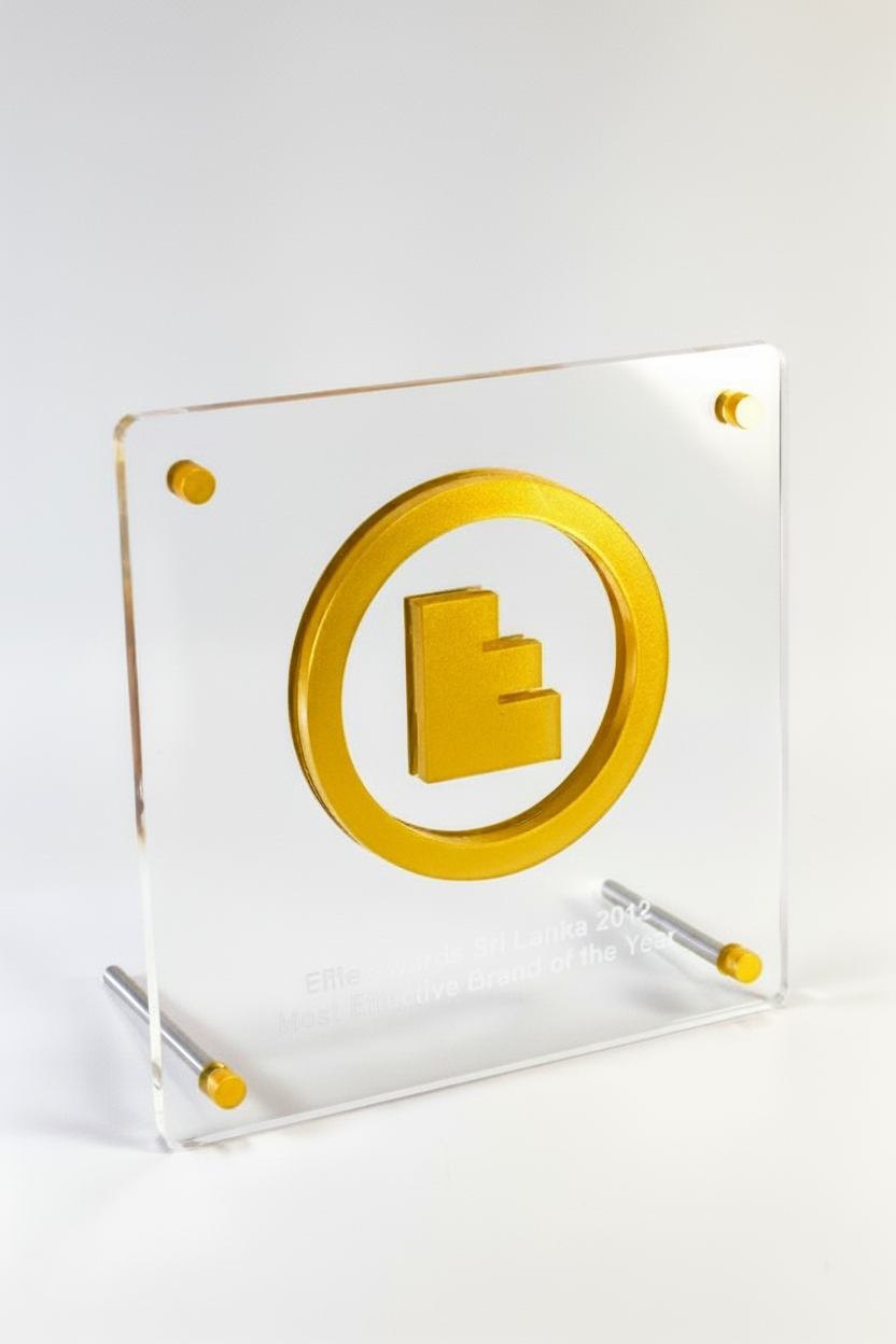 A clear acrylic award plaque featuring a gold logo of a stylized building or steps, with small gold decorative accents on the corners, possibly indicating it was awarded in Sri Lanka in 2012.