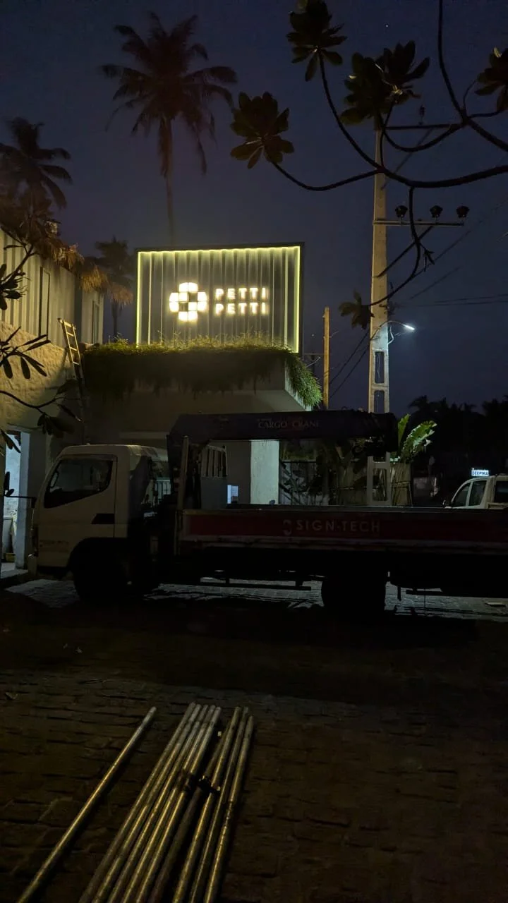Nighttime scene with palm trees, a building featuring a illuminated sign that says 'PETTI PETTI,' and a truck parked on the street. There are some metal poles on the ground in the foreground.