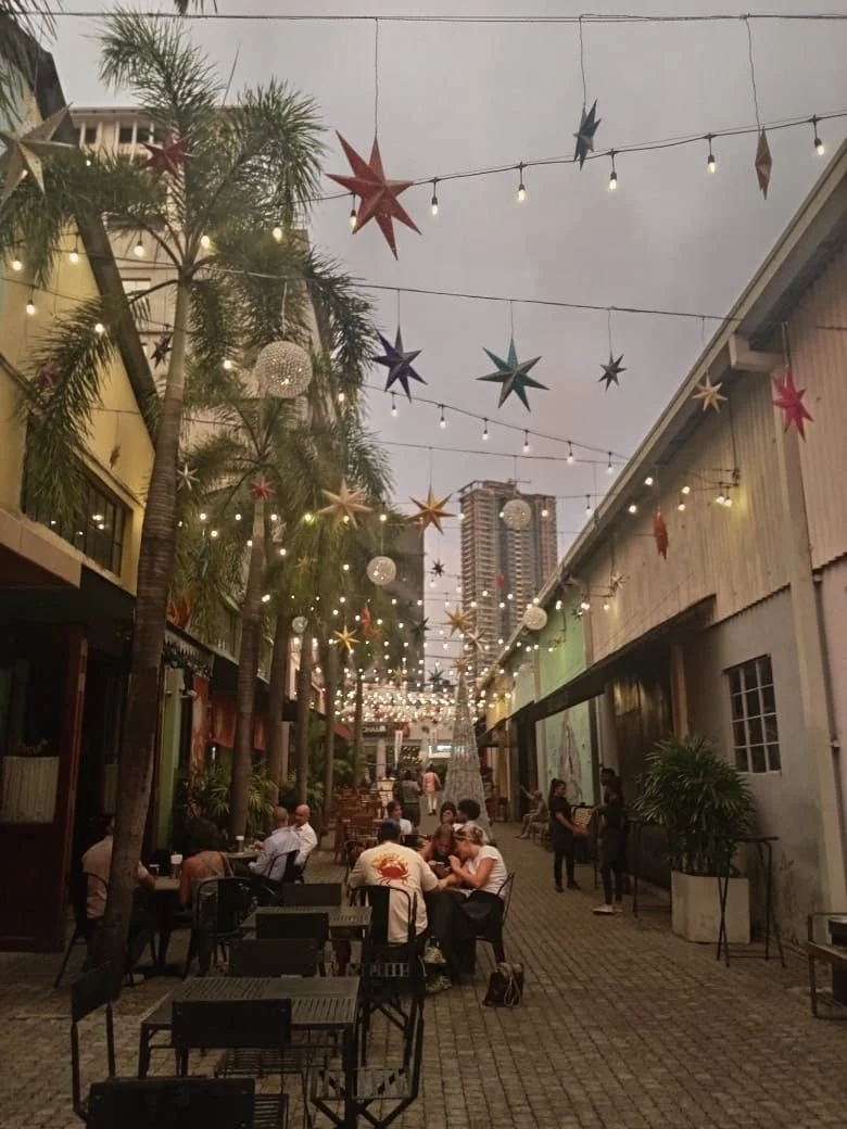Outdoor dining area decorated with hanging star and string lights, palm trees, and a mix of tables and chairs, with people dining and socializing.