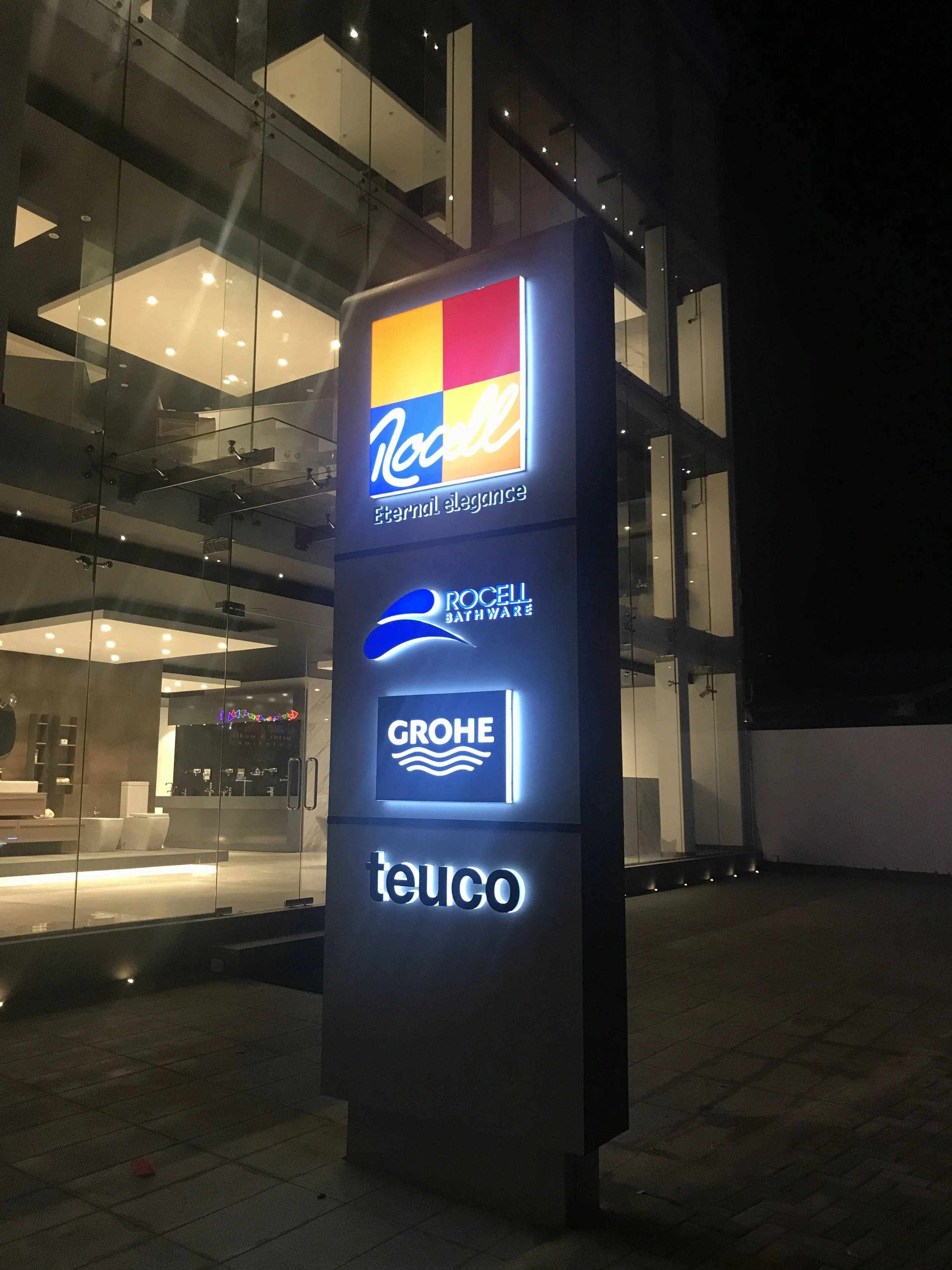 Nighttime exterior view of a modern glass building with illuminated signs. The signs include 'Nacll', 'Rocell Bathware', 'GROHE', and 'teuco'.