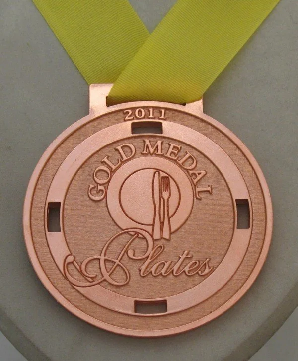 A bronze medal with a yellow ribbon attached, featuring the text '2011 Gold Medal Plates' and an engraved fork and plate emblem.