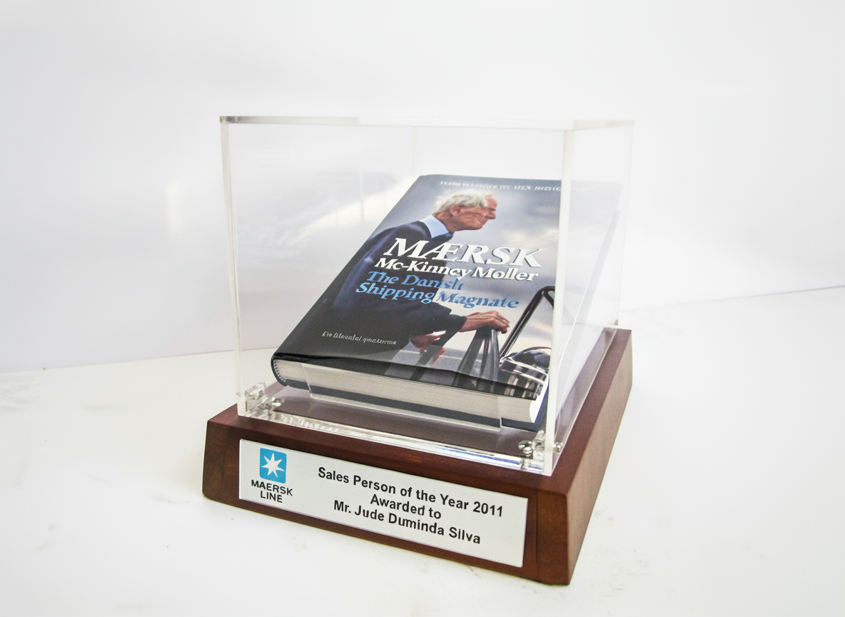 A white trophy case with a clear front, displaying a large hardcover book titled "Maersk The Daring Shipping Magnate" by McKinney Moller. The case is placed on a wooden base with a plaque that reads "Sales Person of the Year 2011 Awarded to Mr. Jude 