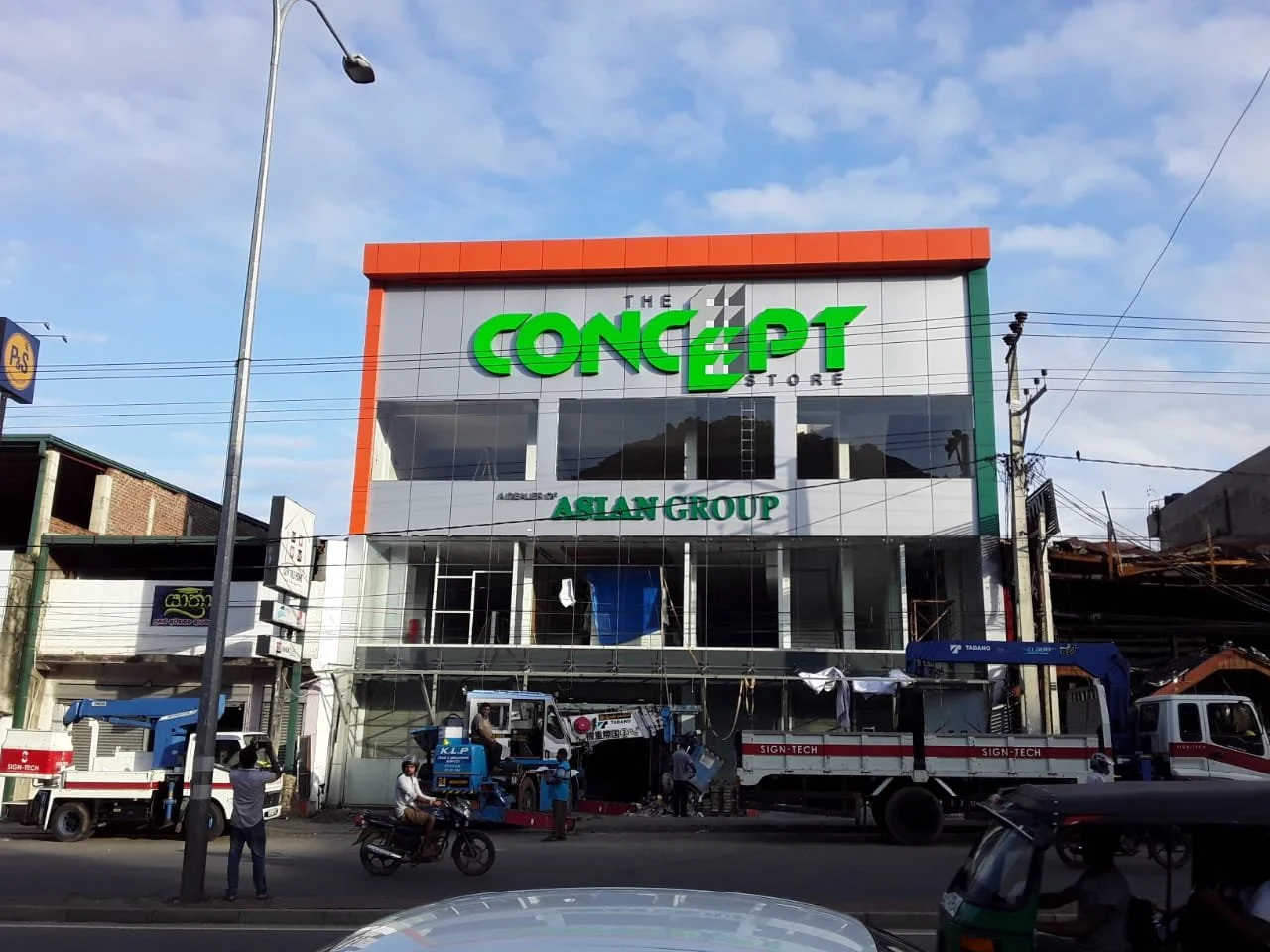 A building under construction with a sign reading 'The Concept Store' and 'Asian Group'. Construction vehicles and workers are present on the street.