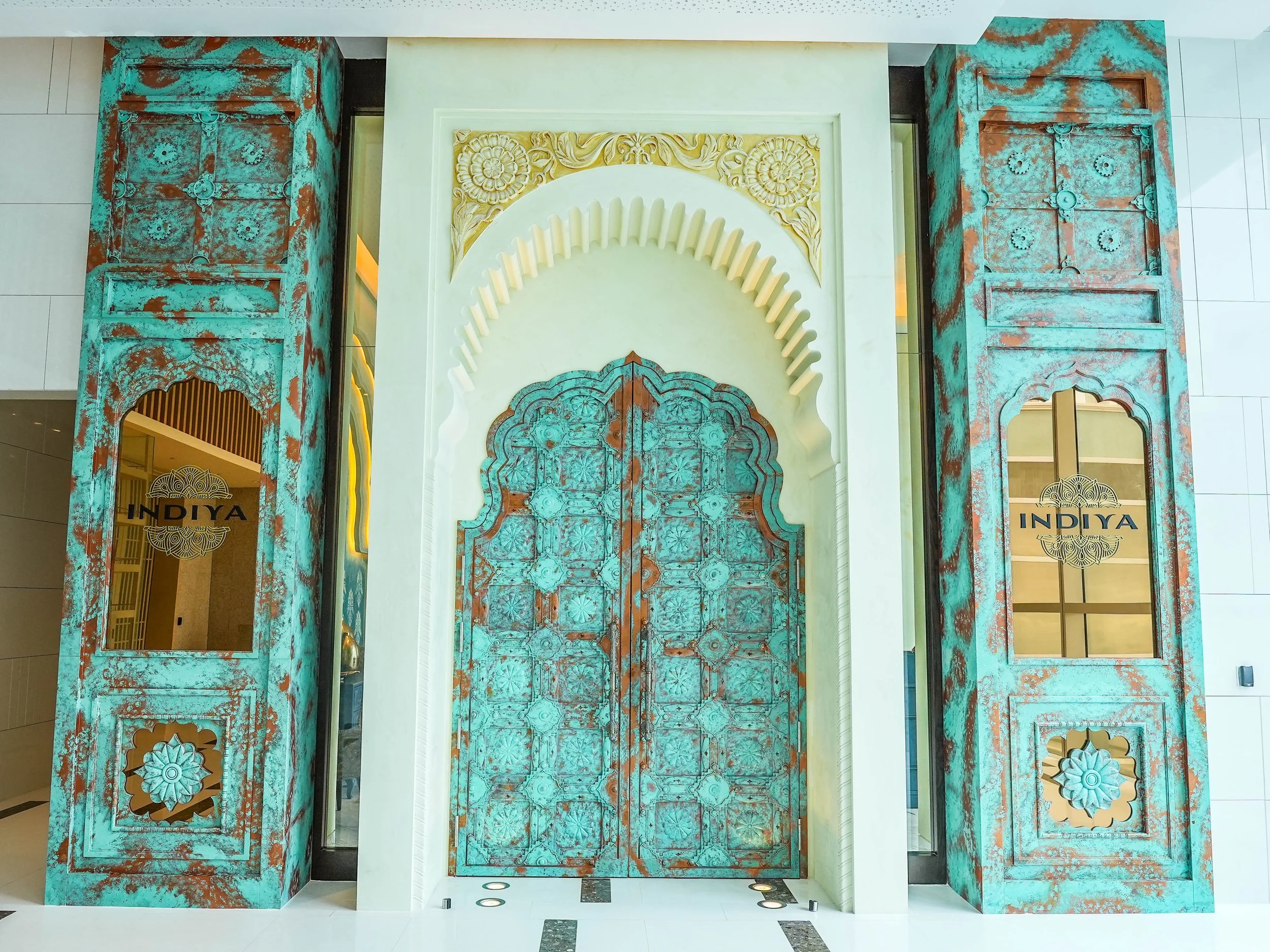 Decorative entryway with teal and rust-colored distressed metal panels, intricate carved stone arch, and golden accents for Indiya restaurant.