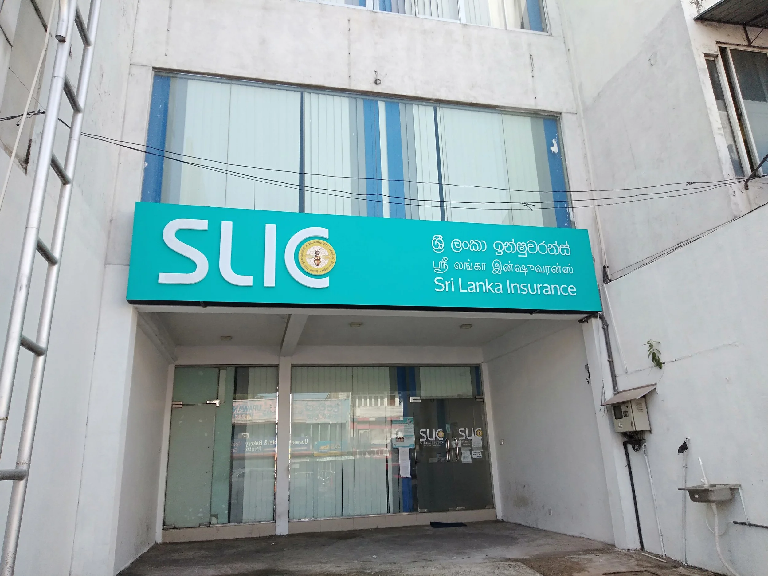 Facade of a building with a large blue sign reading 'SIC' and 'Sri Lanka Insurance' in multiple languages including Sinhala, Tamil, and English, above the entrance, with glass doors and windows, an aluminum ladder on the left side, and power lines ac