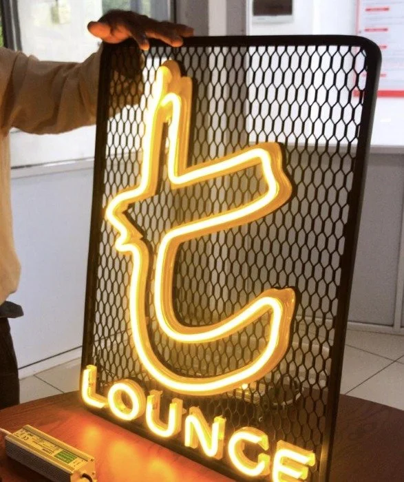Neon sign in the shape of a thumbs-up with the word "LOUNGE" below, mounted on a black mesh panel.