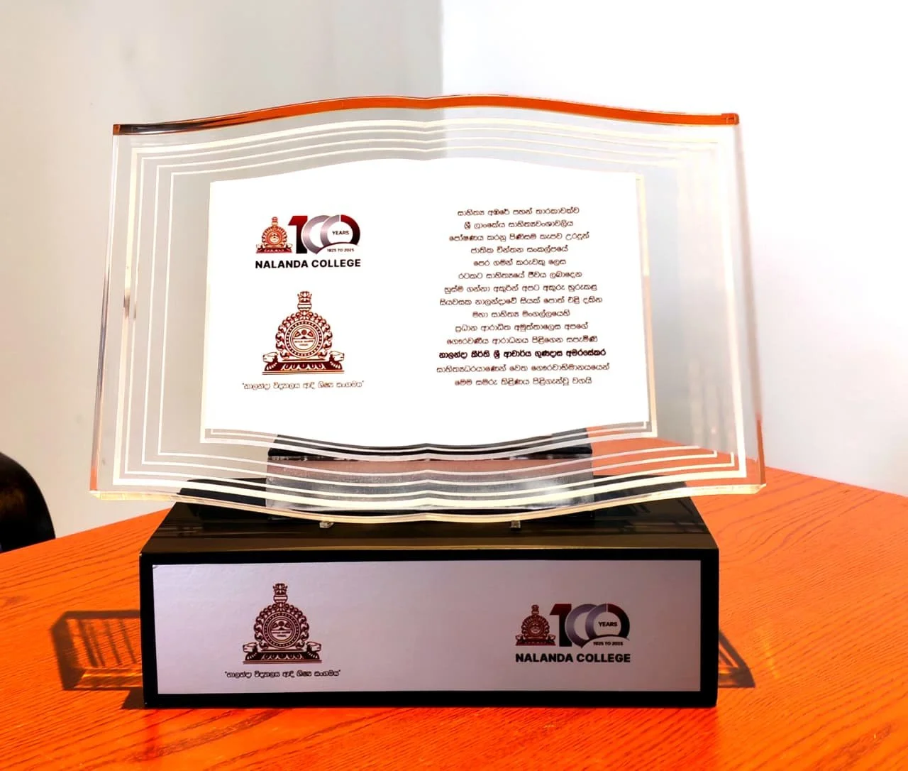 A glass award plaque with the Nalanda College logo, a 100-year anniversary logo, and Sinhala text, sitting on a black base with a label featuring the college logo and anniversary logo, placed on an orange wooden table.