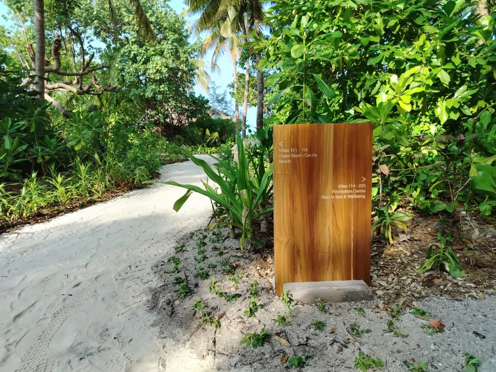 A sandy pathway surrounded by green tropical plants and trees, with a wooden directional sign showing villa and recreation center locations.
