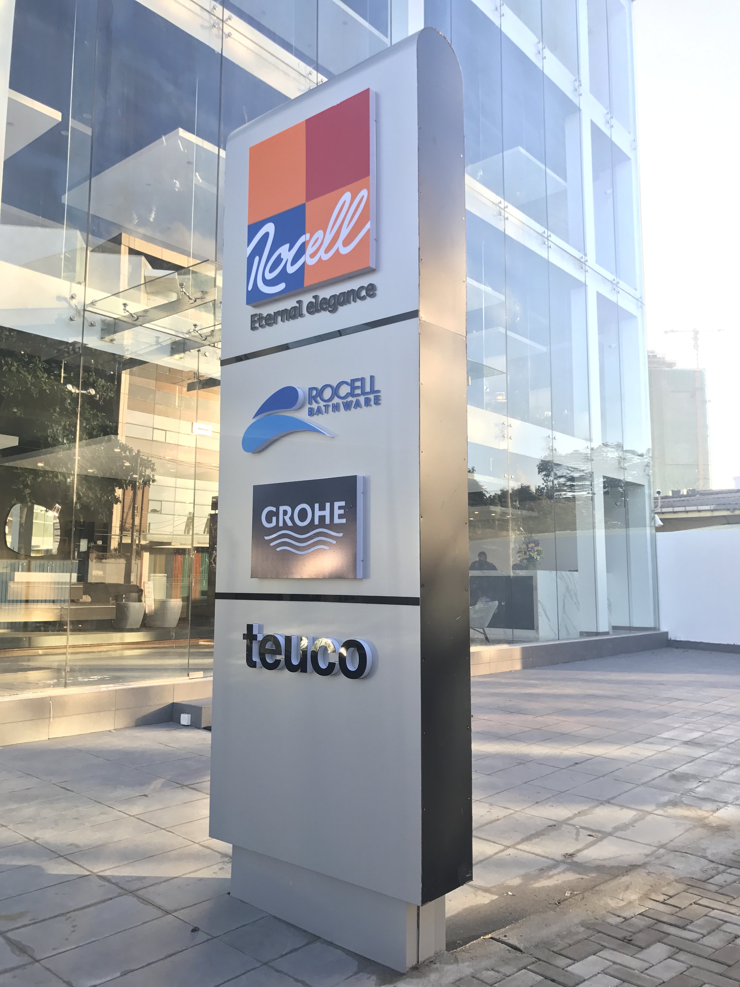 Signboard outside a modern building displaying logos of Rocell, Grohe, and Teuco with reflective glass windows in the background.