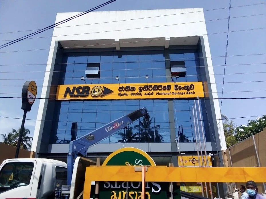 Modern glass-fronted building with a yellow sign for National Savings Bank (NSB) in Sinhala and English, a crane nearby, and a partial view of a bus stop sign and people in masks.